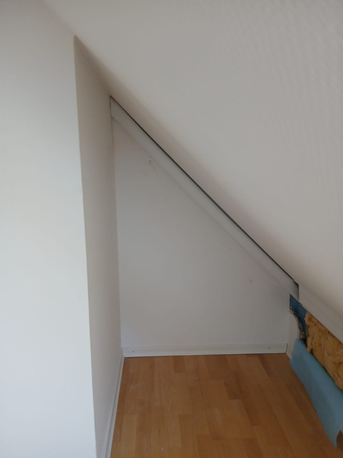 Schräge Nische mit weißer Verkleidung und sichtbarer Rohrleitung in einem Raum mit Laminatboden.
